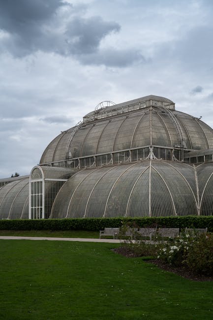 Photograph of the large glasshouses at Kew Gardens, featuring a domed, multi-tiered Victorian-style conservatory with curved glass panels supported by metal framework. In the foreground, there is a we