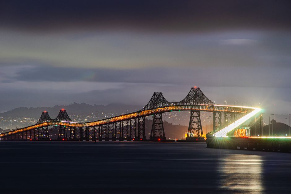 A large steel suspension bridge extends across a body of water beneath a sky with a full moon partially obscured by a thick layer of clouds. The bridge's towers and cables are clearly visible, with th